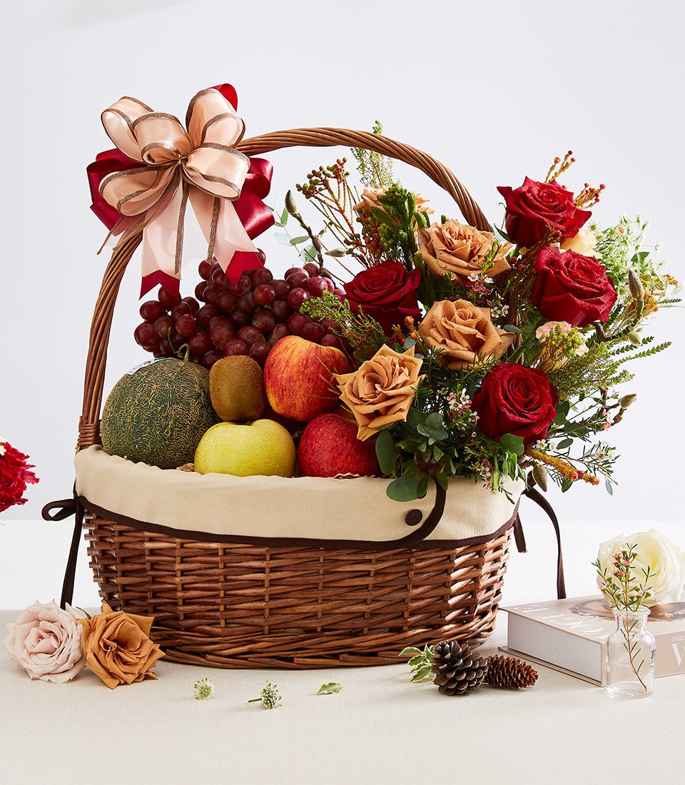 Wicker gift basket with fresh fruits, red and peach roses, and layered pink-red ribbon bow