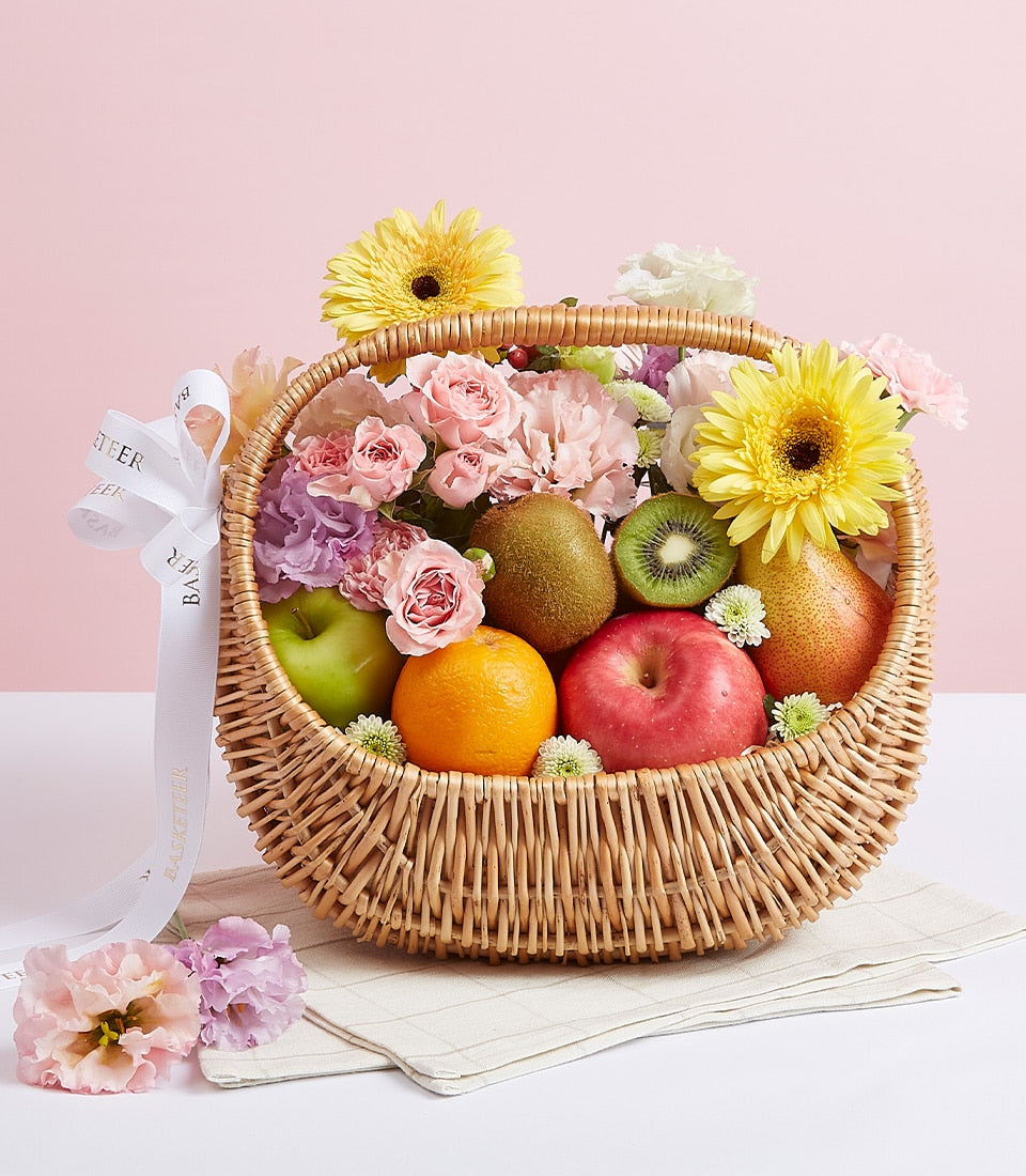 Woven wicker fruit basket with fresh apples, kiwis, and pears, accented with pink roses and yellow gerberas for birthdays