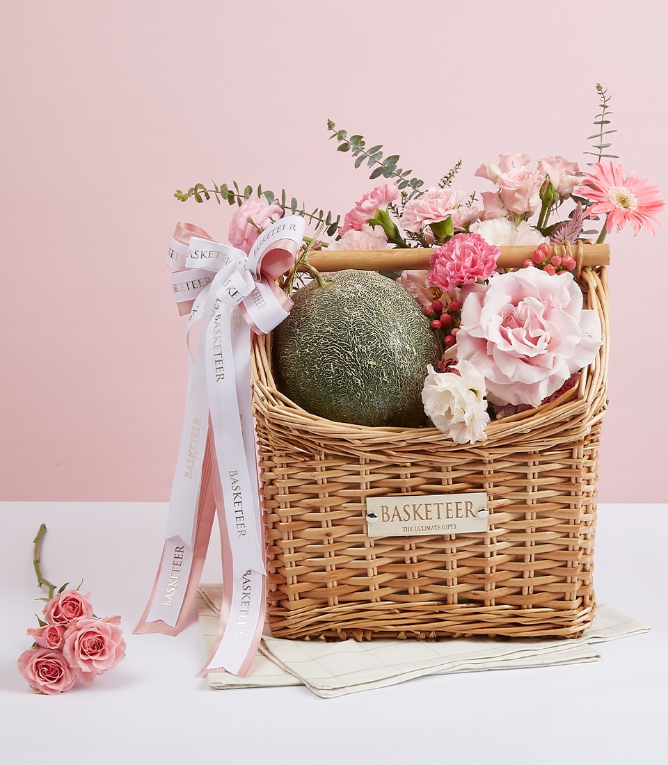 Woven wicker gift basket with fresh cantaloupe melon, pink roses, carnations, and gerbera daisy with white ribbon bow