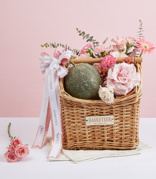 Woven wicker gift basket with fresh cantaloupe melon, pink roses, carnations, and gerbera daisy with white ribbon bow