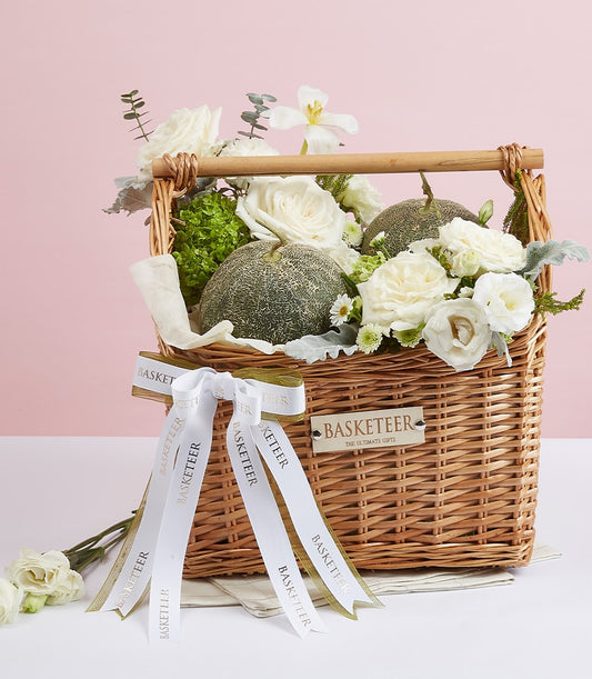 Woven wicker gift basket with fresh green melons, white roses, ranunculus, and premium foliage arrangement