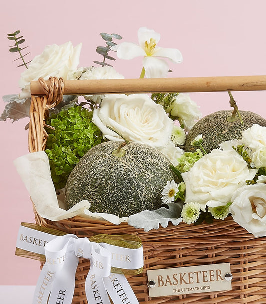 Woven wicker gift basket with fresh green melons, white roses and tulips, eucalyptus, and cream ribbon