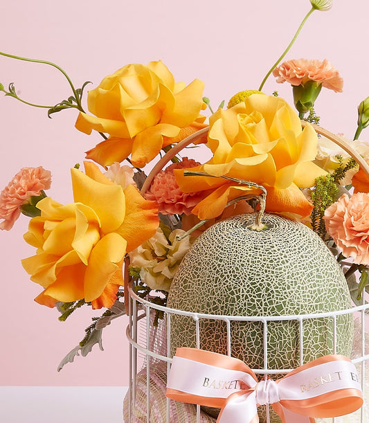 Premium yellow roses and fresh cantaloupe melon in white wire gift basket with orange carnation accents