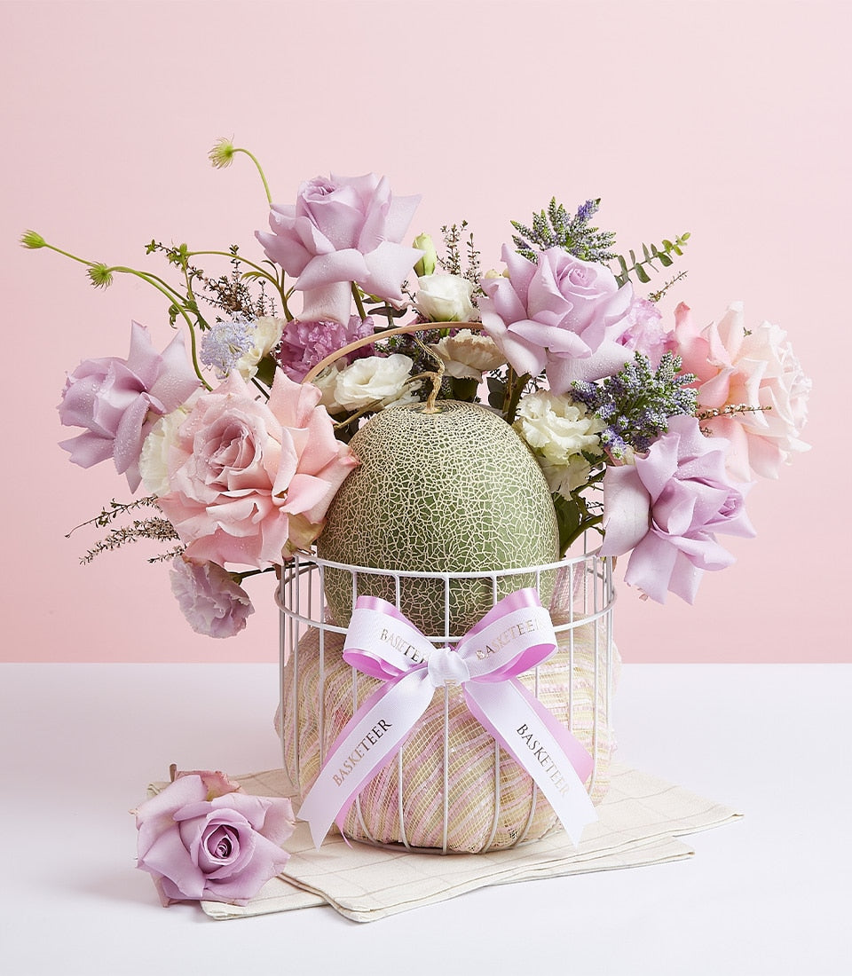Fresh melon fruit and lavender rose bouquet in white wire basket with pink ribbon