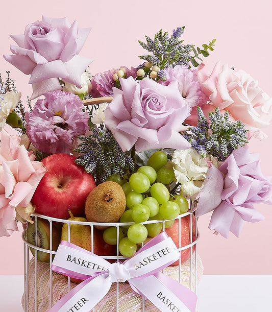 Purple and pink roses with fresh fruit in white wire basket, lavender carnations and kiwis for birthday gifts