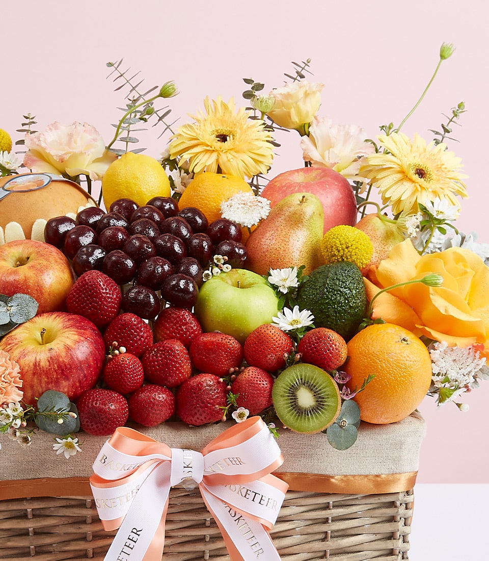 Vibrant mixed fruit gift basket with fresh apples, strawberries, citrus, and yellow gerbera daisies in woven wicker with