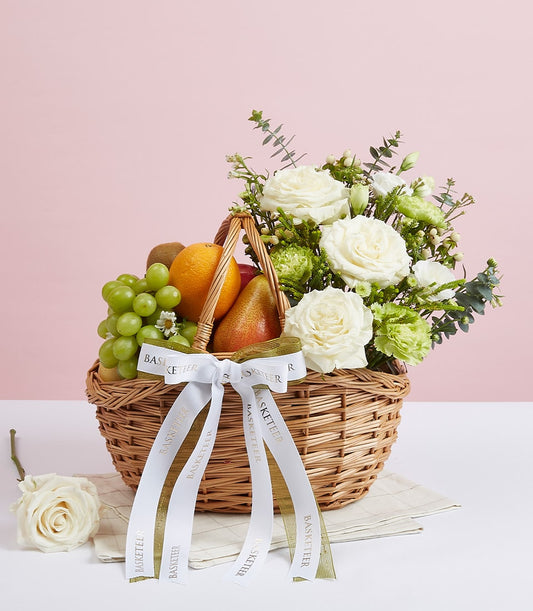 Woven wicker fruit basket with fresh grapes, apple, pear, orange, kiwi and white roses on soft pink background