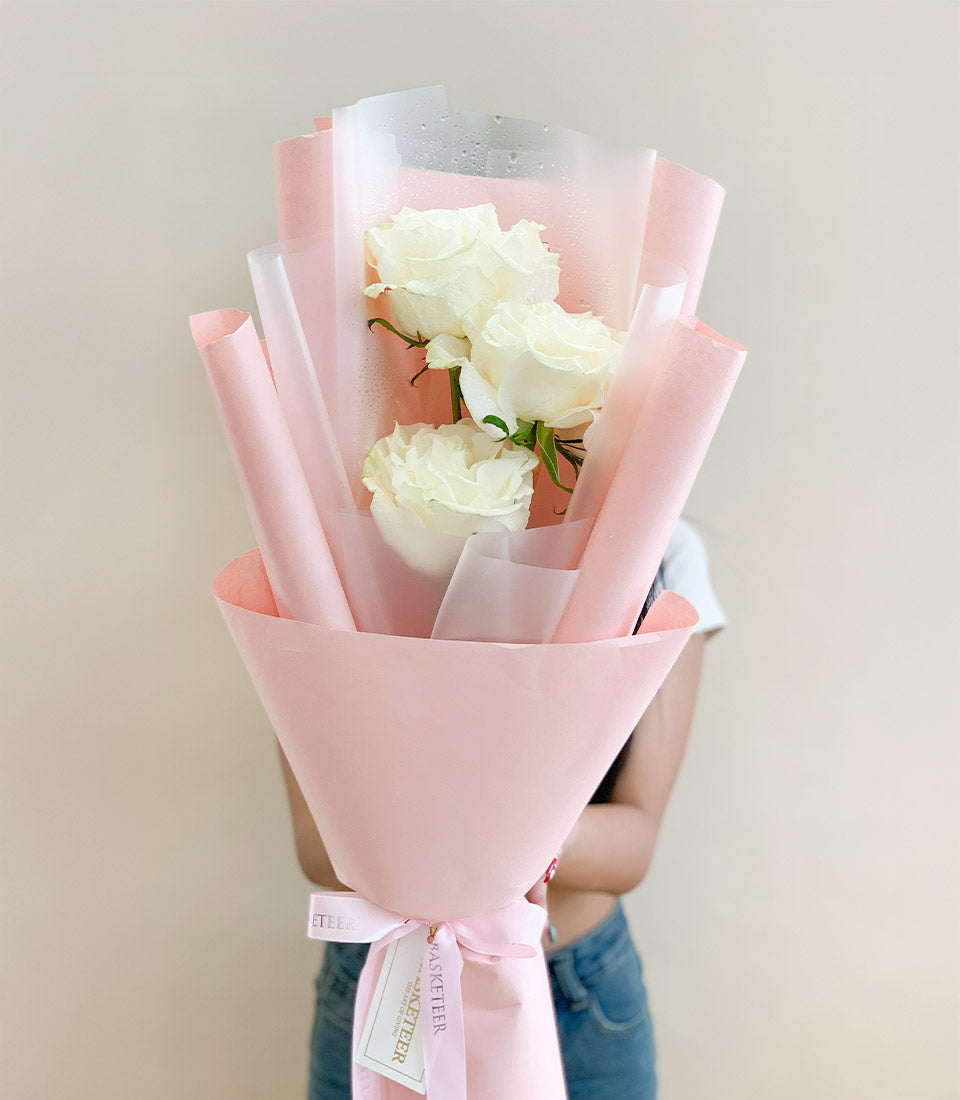 Three fresh white Mondial roses with dewy petals, wrapped in soft pink paper and tied with ribbon