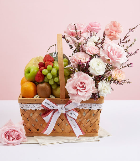 Light wood gift basket with fresh fruit, pink and white flowers, and white lace trim on soft pink backdrop