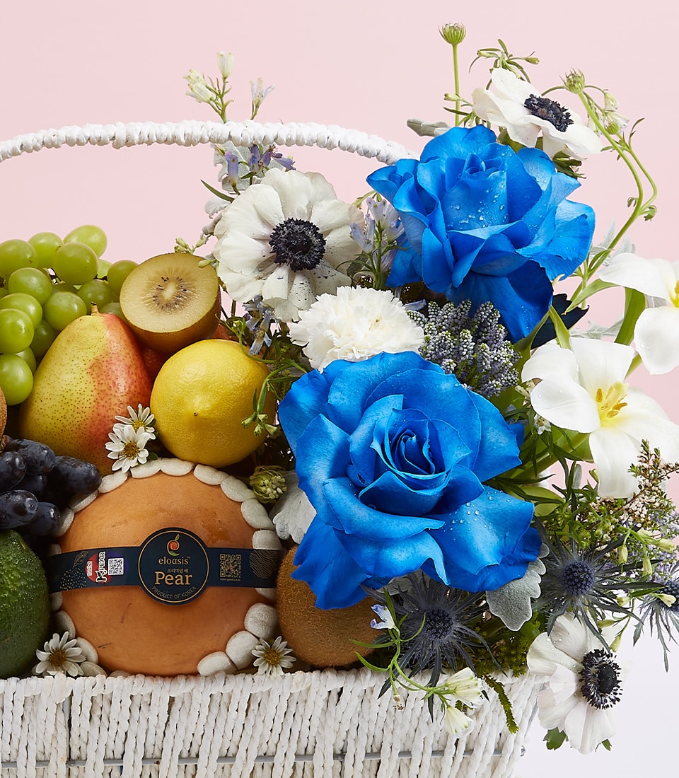 Premium blue roses and fresh fruit selection in white woven basket with white anemones and tropical fruits