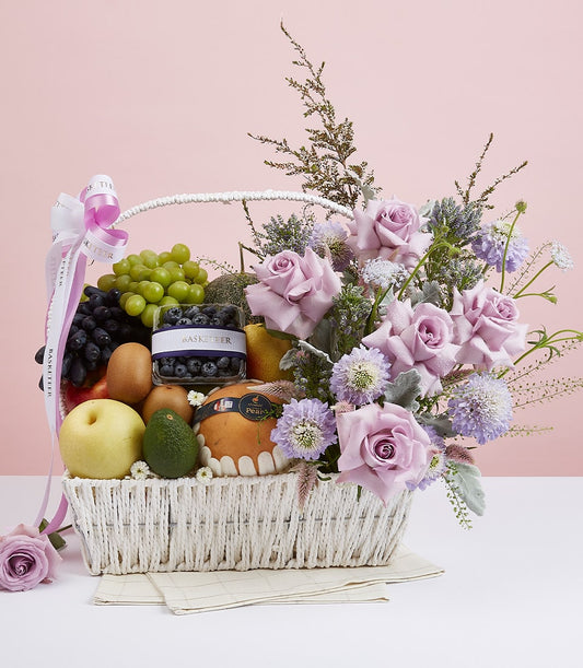 White woven basket with fresh fruits, blueberries, and light purple roses with greenery, white ribbon accent