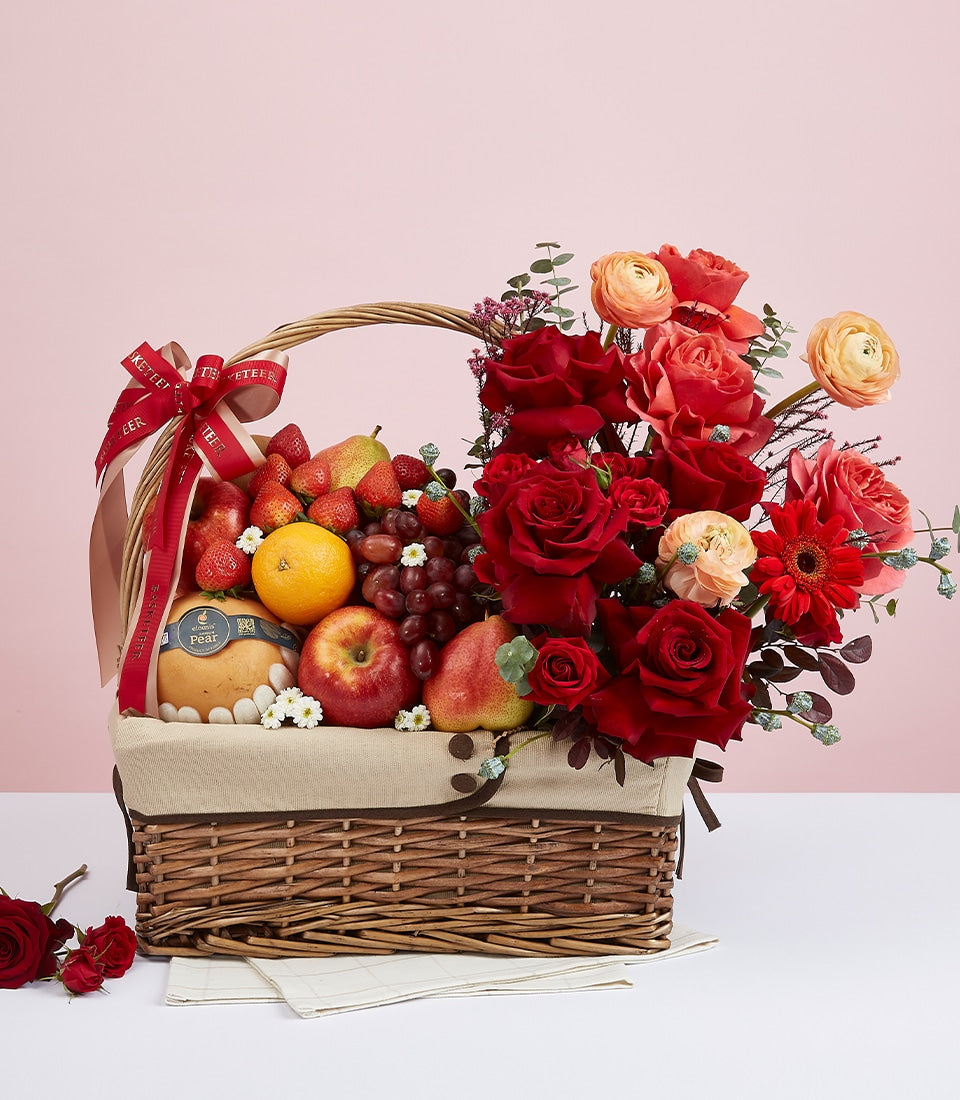 Mixed fresh fruit and premium red roses in woven gift basket with coral ranunculus and gerbera, perfect for celebrations and