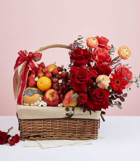 Mixed fresh fruit and premium red roses in woven gift basket with coral ranunculus and gerbera, perfect for celebrations and