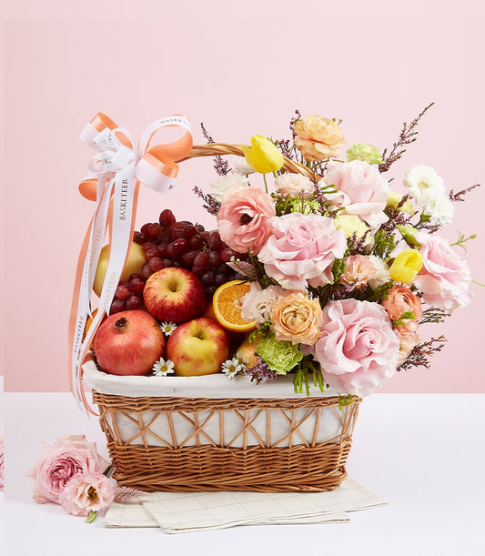 Premium wicker basket with fresh fruit, roses, and daisies in pink, peach, and yellow tones