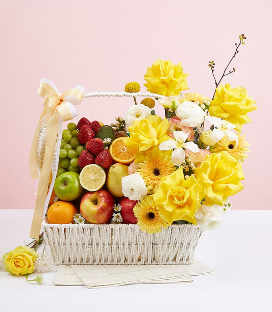 Cream ribbon-tied woven basket with fresh mixed fruits, yellow roses, and white gerbera daisies