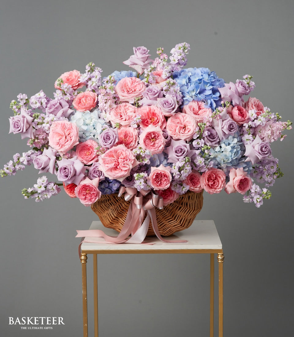 Large woven basket filled with pink and purple roses, blue hydrangeas, and lavender stock with pink satin ribbon
