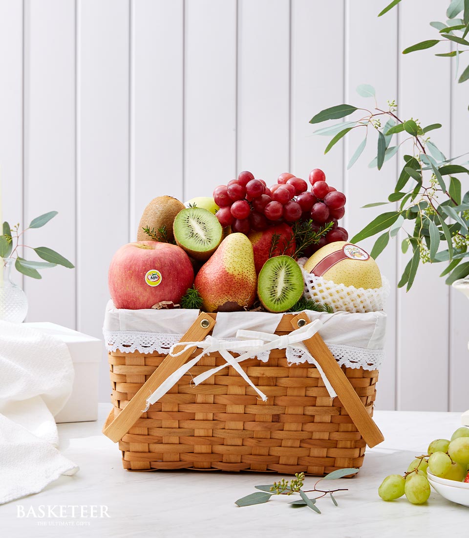 Rustic woven fruit basket with red apples, pears, kiwis, grapes, white lace liner and ribbon bow