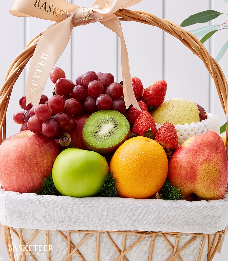 Wicker fruit basket with fresh grapes, apples, kiwi, strawberries, orange and pear, white fabric liner, beige ribbon
