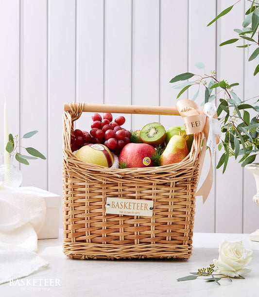 Woven wicker fruit basket with fresh red grapes, apples, pear, and kiwi, tied with beige ribbon