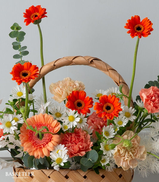 Vibrant orange gerbera daisies and peach carnations in woven rattan basket with eucalyptus greenery