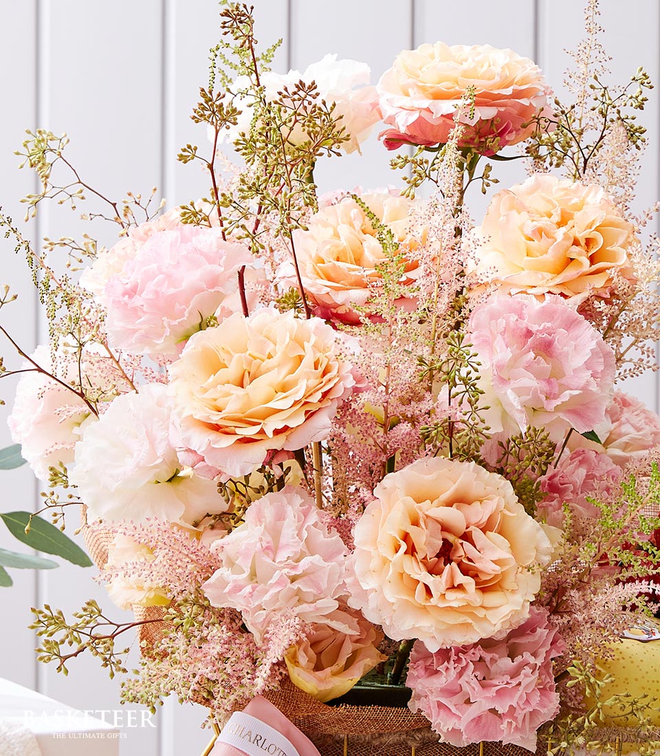 Soft peach and pink garden roses with carnations in burlap-wrapped fruit basket with white ribbon