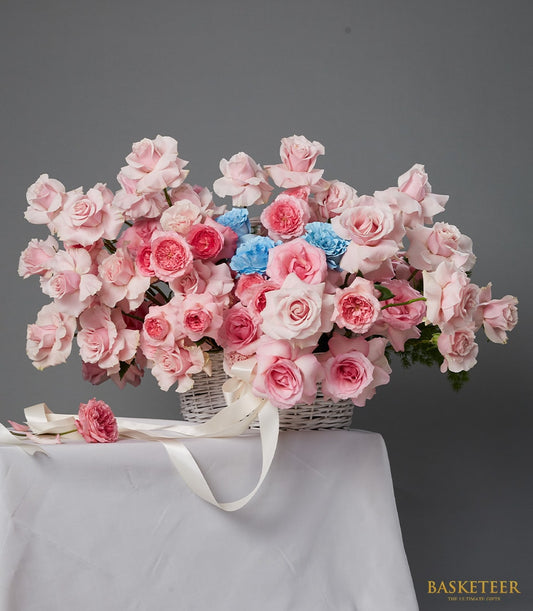Blush pink roses with blue accents in white woven basket, anniversary flower gift