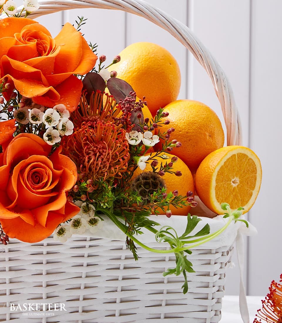 Premium orange roses and fresh oranges in white woven basket with protea and seasonal foliage
