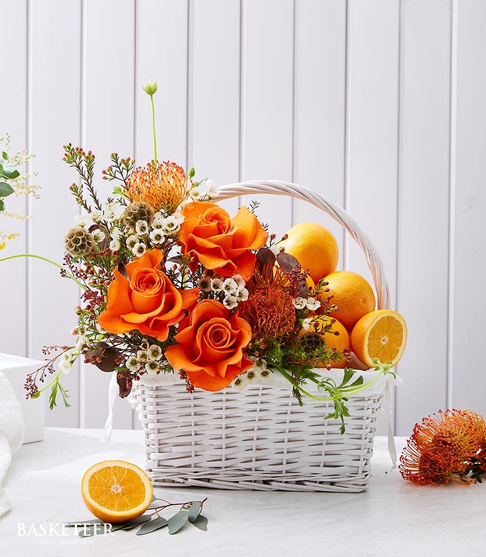 Premium orange roses and fresh citrus in white wicker harvest basket with protea accents