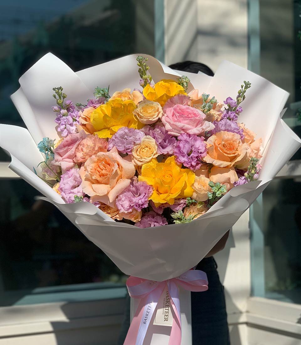 Soft Seasons Harmony graduation bouquet with yellow, pink, peach roses and purple accents, white wrapped