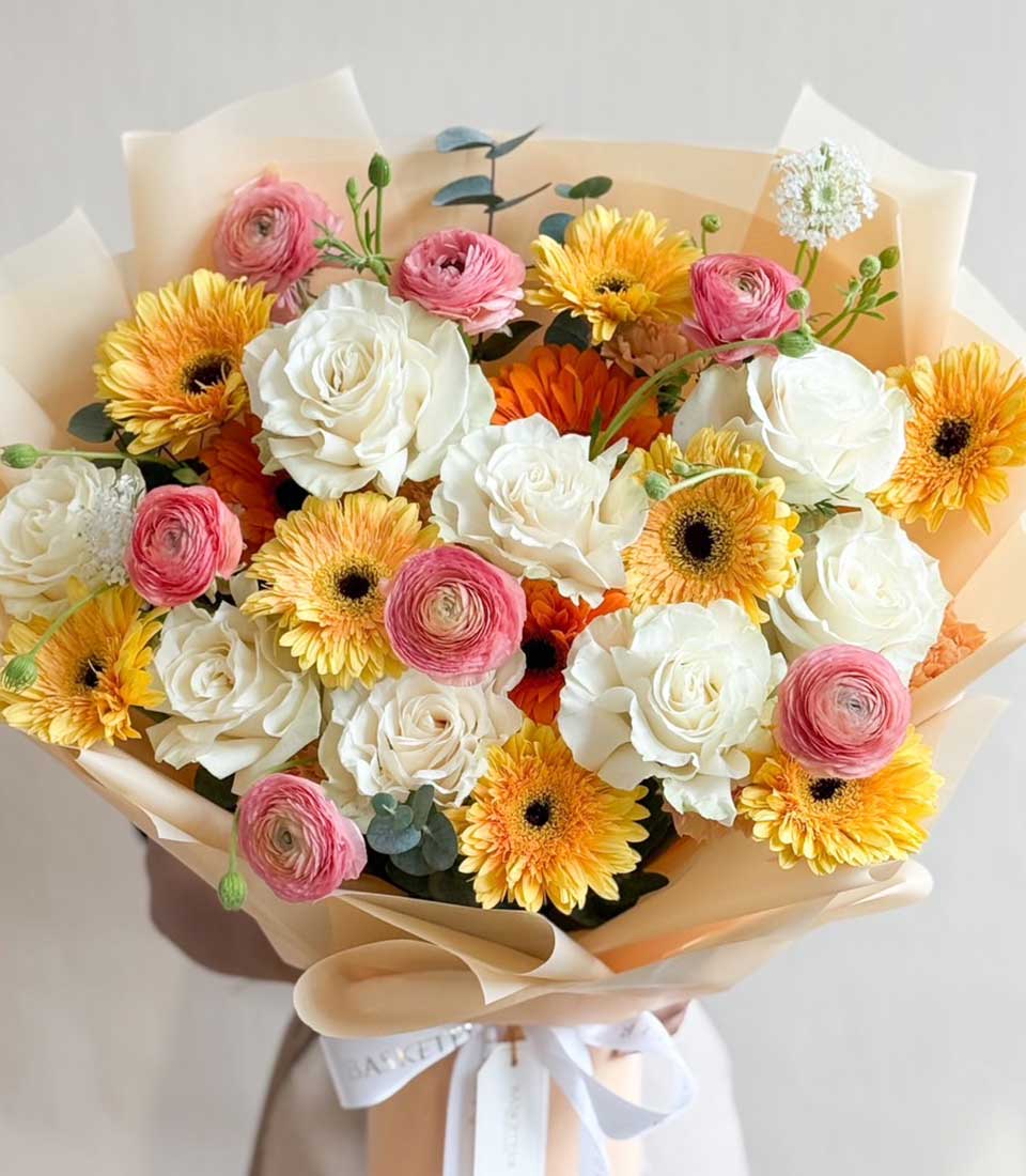 Fresh white roses, pink ranunculus, and yellow gerberas in beige paper wrap with satin ribbon