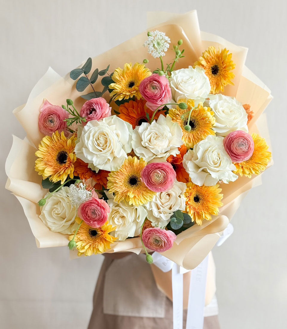 Sunshine & Blush bouquet with white roses, yellow gerbera daisies, pink ranunculus and eucalyptus, wrapped in cream paper