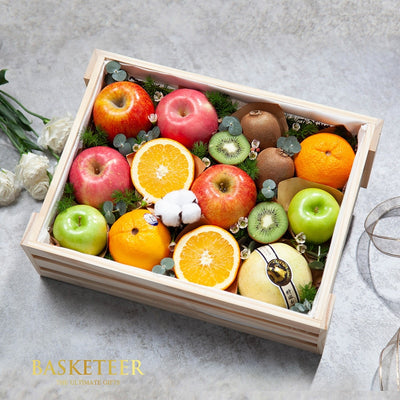 Wooden crate of fresh assorted fruits with apples, oranges, kiwis, and pear nestled in greenery and white flowers