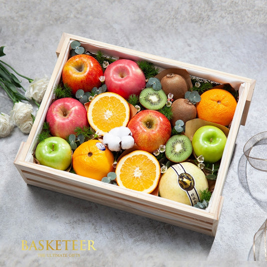 Wooden crate of fresh assorted fruits with apples, oranges, kiwis, and pear nestled in greenery and white flowers