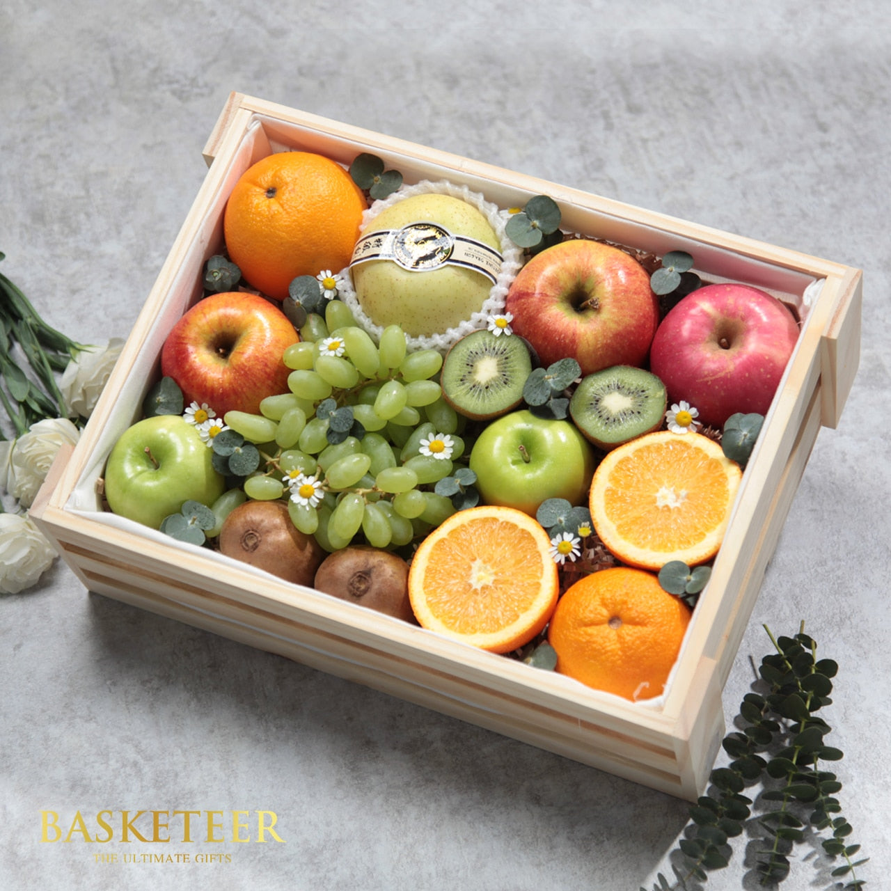 Wooden gift crate filled with fresh fruit, apples, pears, grapes, kiwis, and citrus with white flowers and eucalyptus