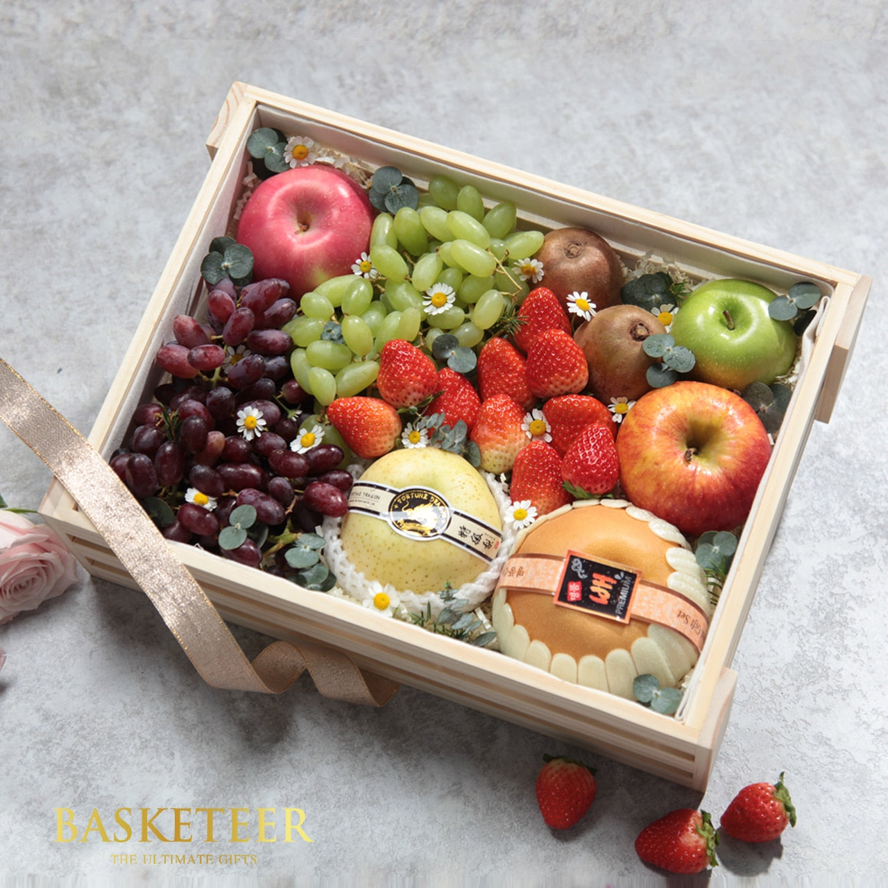 Wooden crate filled with fresh grapes, apples, kiwis, strawberries, and wrapped pears with white flowers and gold ribbon