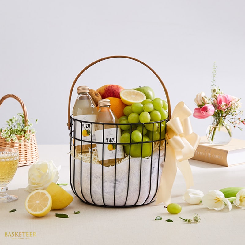 Premium wellness fruit basket with fresh apples, grapes, citrus, and yuzu beverages in black wire basket with yellow ribbon