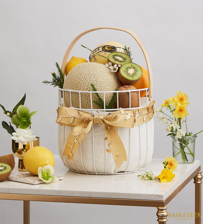 Orchard Fresh Fruit Bounty white wire basket with cantaloupes, kiwis, lemons, oranges, and fresh florals