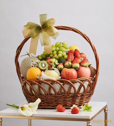 Woven dark brown fruit basket with gold-green ribbon bow, filled with fresh grapes, strawberries, apples, cantaloupe, and