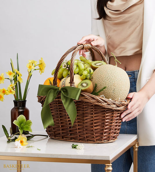 Luxury woven wicker fruit basket with green grapes, cantaloupe, kiwis, pear, and orange tied with dark green satin ribbon