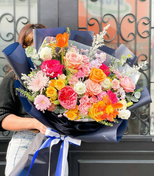 Vibrant mixed flower bouquet with orange tulips, pink roses, and red peonies tied in dark blue paper with white striped