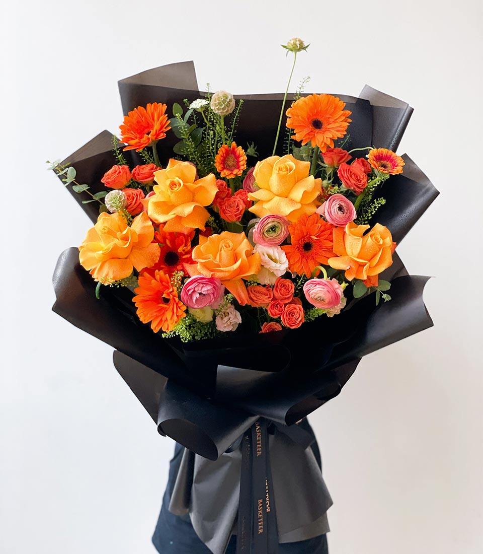 Vibrant orange and yellow gerbera daisy bouquet with roses and ranunculus, wrapped in black paper with gold ribbon