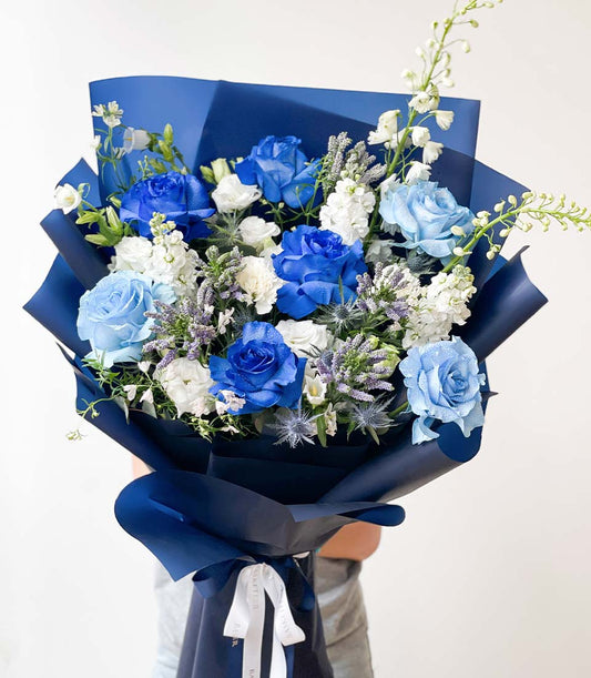 Vibrant blue and white fresh flower bouquet with roses, carnations, and thistle wrapped in dark blue paper