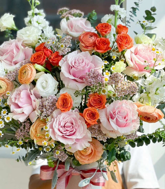 Vibrant mixed rose vase with orange, pink, and red blooms, white stock flowers, and delicate purple filler in white