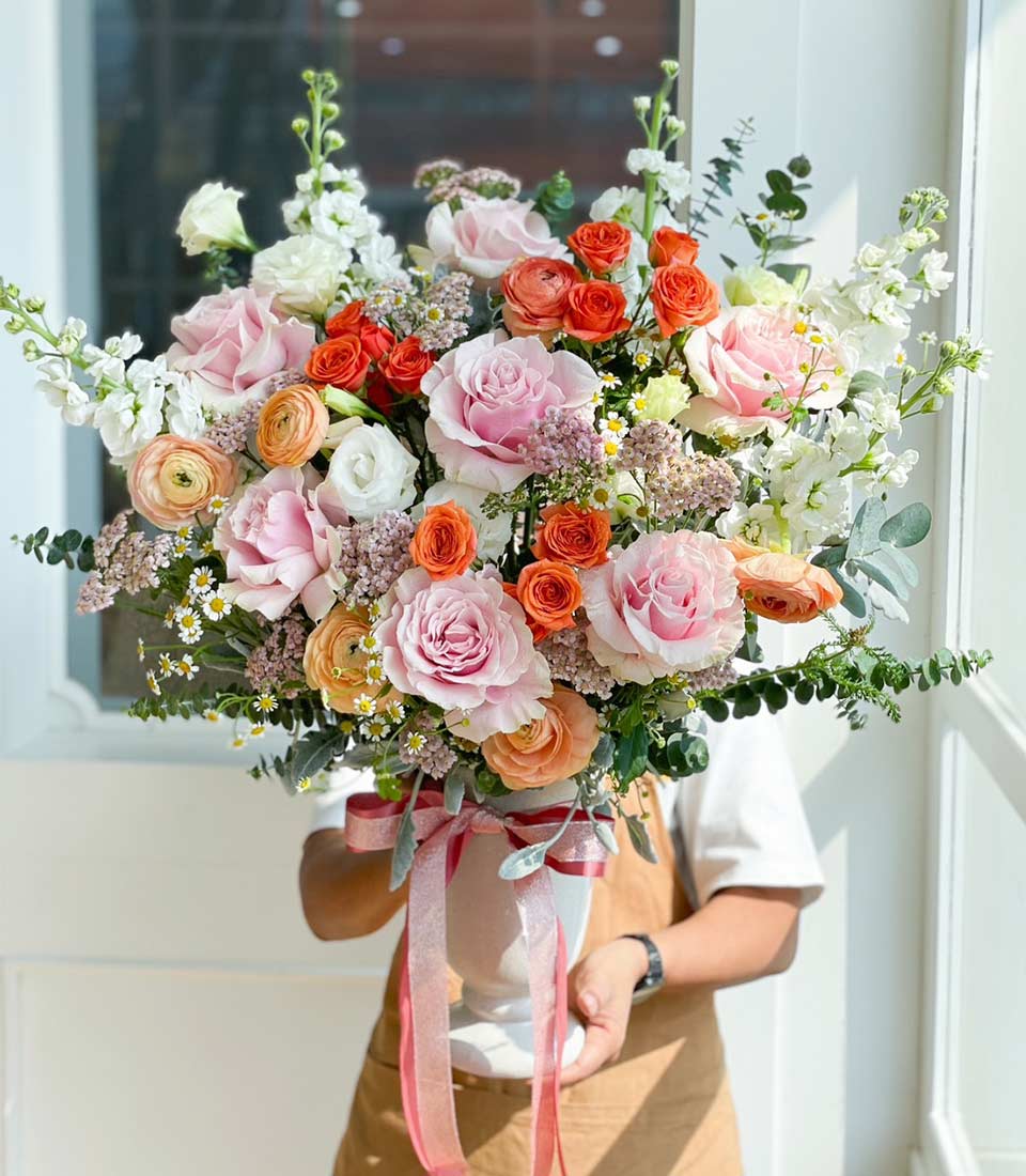 Vibrant bouquet of pink, orange and white roses with stock flowers in white fluted vase, tied with striped ribbon