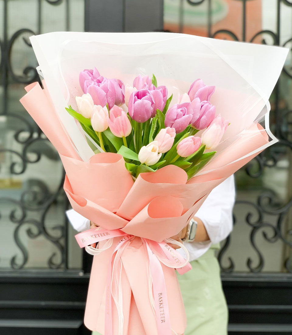 Pink and white tulip bouquet with purple-pink accents, wrapped in layered light pink paper with Basketeer ribbon bow for
