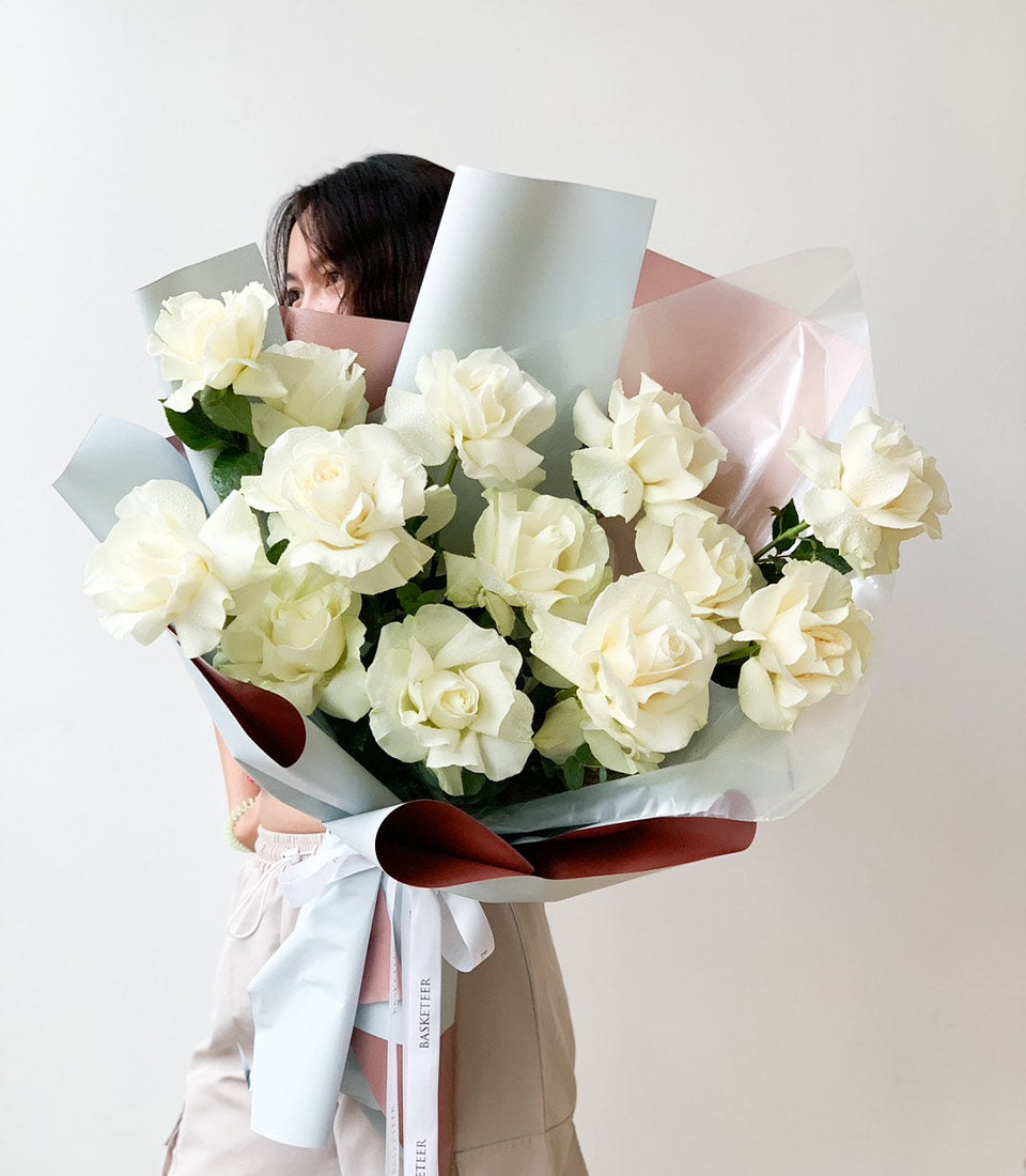Large white rose bouquet with water droplets, wrapped in layered blue, pink, and brown paper with white ribbon