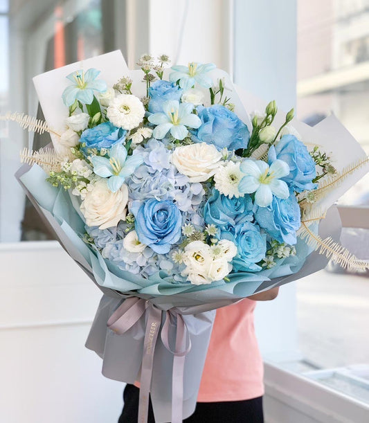 Serene blue and white rose bouquet with hydrangeas and lilies, wrapped in layered blue paper and grey ribbon