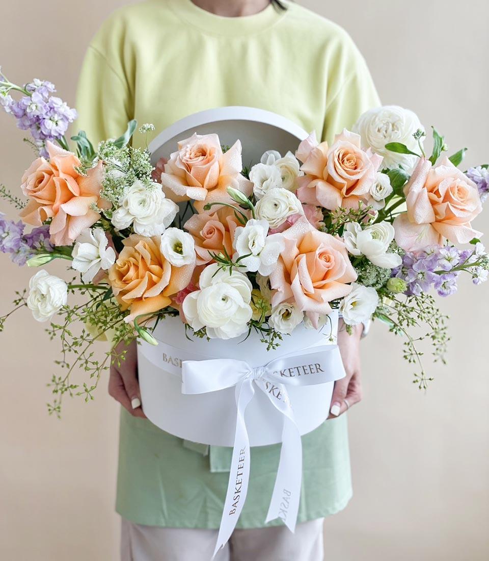 Large white hatbox filled with fresh peach roses, white ranunculus, and purple stock flowers tied with satin ribbon, ideal