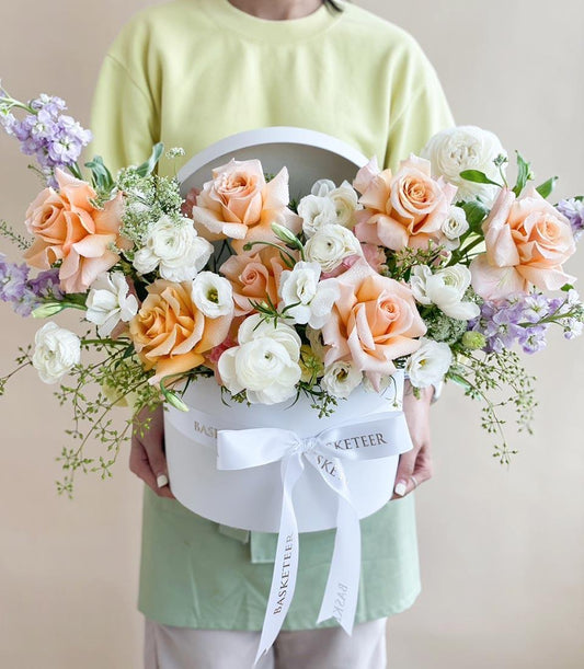 Large white hatbox filled with fresh peach roses, white ranunculus, and purple stock flowers tied with satin ribbon, ideal