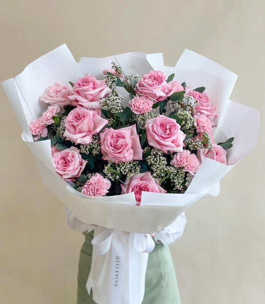 Pink roses and carnations fresh flower bouquet with white filler and green foliage, wrapped in white paper with satin ribbon